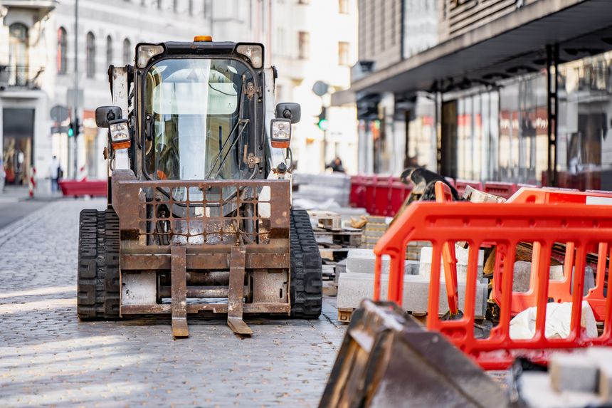 Compact tracked skid steer loader with forklift attachment parked on a city street pavement, representing municipal urban infrastructure construction