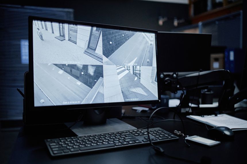 Computer monitor displaying multiple security camera feeds showing empty urban streets, keyboard and mouse placed on desk, surveillance equipment used for monitoring public areas