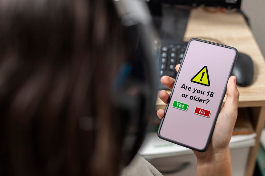 An unrecognizable female teen is holding a mobile phone with legal age verification displaying on its screen