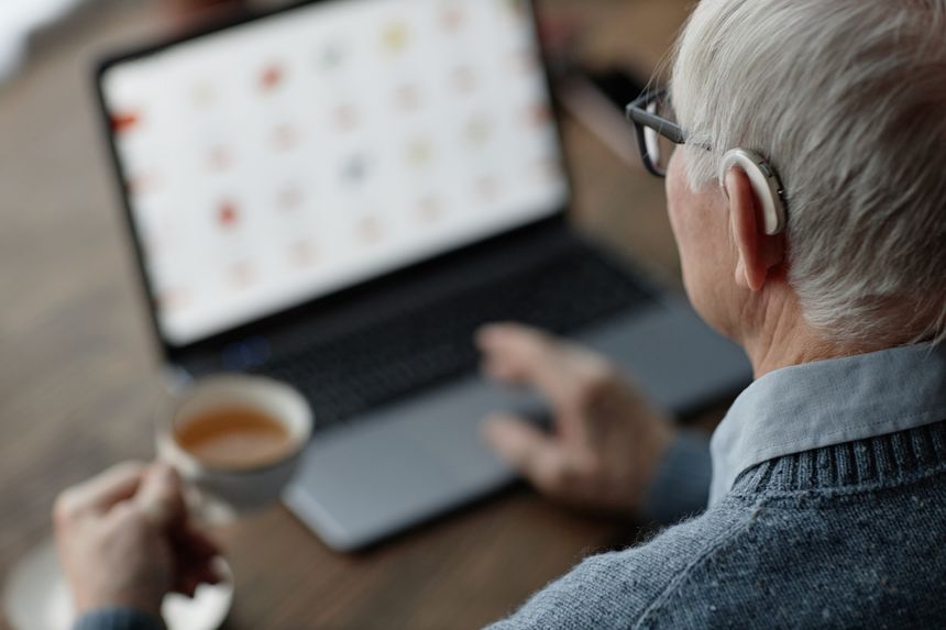 Over shoulder shot of elderly man wearing hearing aid behind ear working on laptop at home while using assistive technology, copy space
