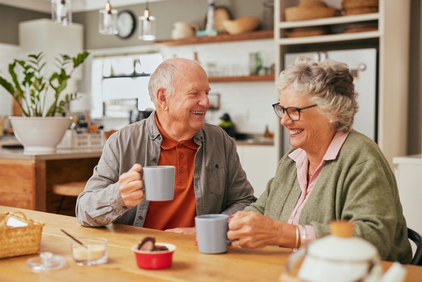 Smiling old couple sitting at table and sharing coffee in their cozy kitchen. Elder man and retired woman laughing together while enjoying a cup of coffee indoors. Relaxed aged couple drinking a cup of tea during a conversation at home while smiling together.
