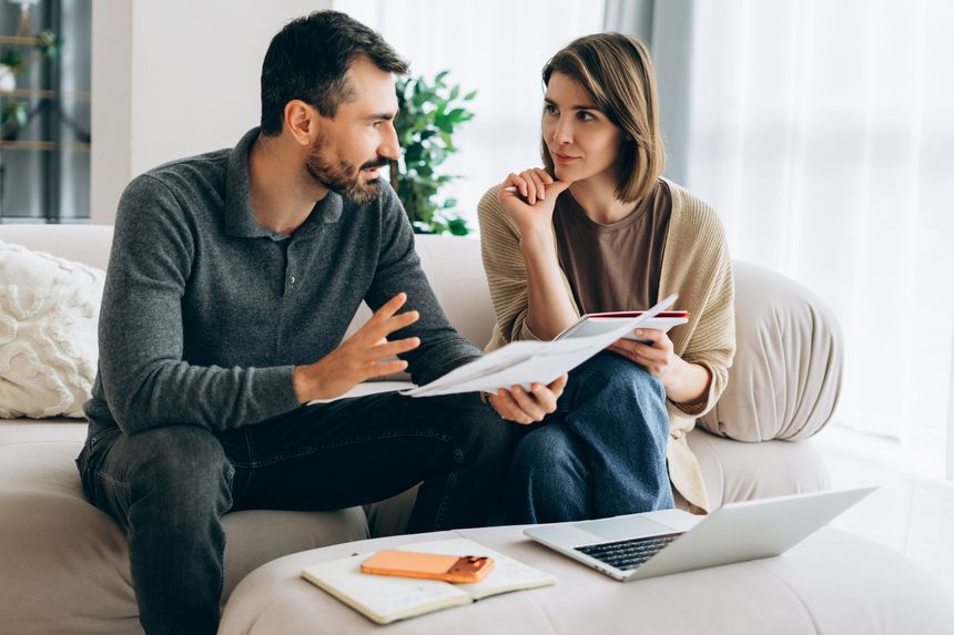 Couple sitting on a couch, seriously discussing financial documents and bills, planning their household budget, and managing expenses at home with a laptop.