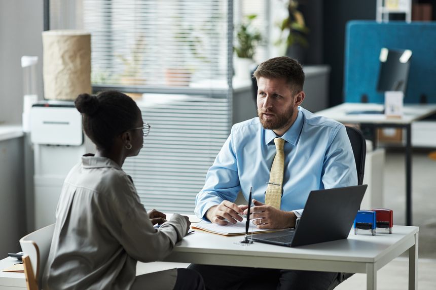 Caucasian male consultant sitting at desk talking with young Black woman in bank office, both engaged in business discussion with documents and laptop on table, professional setting