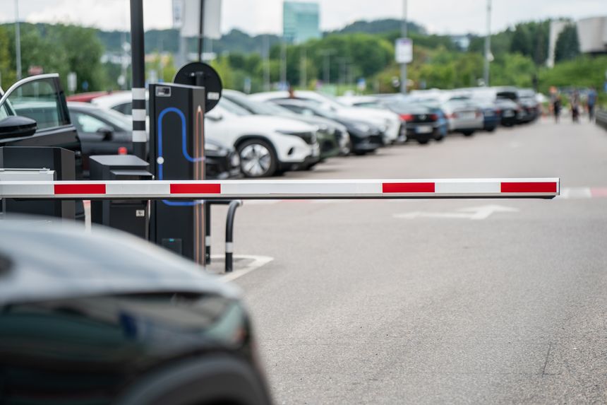 A suburban automated parking lot barrier gate is shown partially lowered, controlling vehicle access. The background features a line of cars parked in neat rows, with lush green trees and buildings visible in the distance under a cloudy sky, suggesting a well-maintained and organized public or private parking facility.