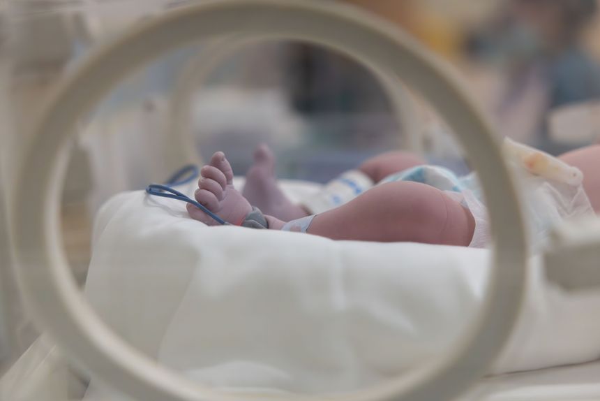 A baby's feet are visible in a hospital incubator. The baby is wrapped in a blanket and has a blue strap around its foot. The baby is in a hospital setting, and the image conveys a sense of care