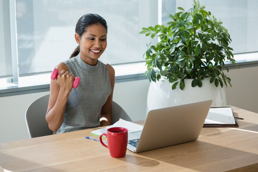 Smiling executive exercising with dumbbells while working on laptop in office