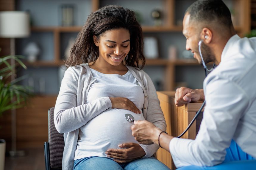 Pregnant millennial black woman sitting on chair at clinic, touching her big tummy and smiling, visiting doctor, arab man gynecologist checking baby heart rate with stethoscope, copy space