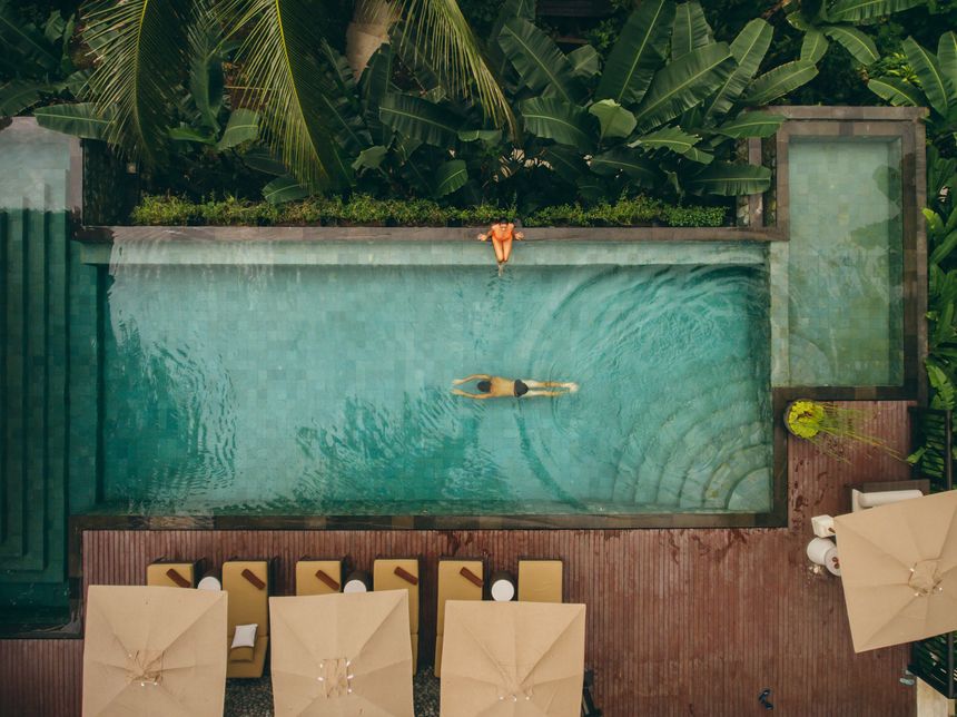 Aerial shot of woman sitting at the poolside with man swimming in pool. Couple enjoying holiday at luxury resort.