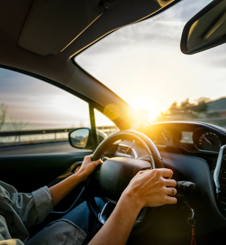 Women's hands on the steering wheel of a car. Woman driving a car at sunset on the seashore. Auto travel concept