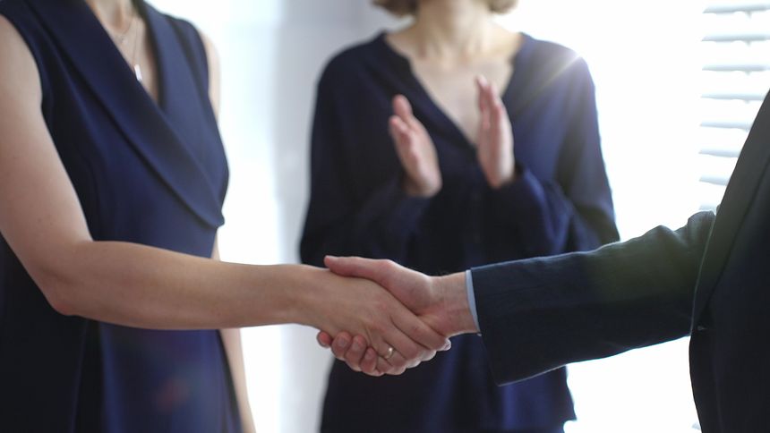 Business people are shaking hands, confirming a successful agreement and strong partnership, as a colleague claps in approval in bright office with sun shines.