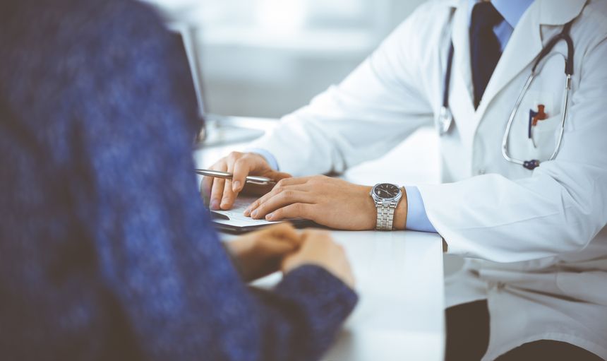 Unknown young woman patient discuss the results of her medical tests with a doctor, while sitting at the desk in a hospital office. Physician using clipboard for filling up medication history records. Perfect medical service in clinic. Medicine and healthcare concepts.