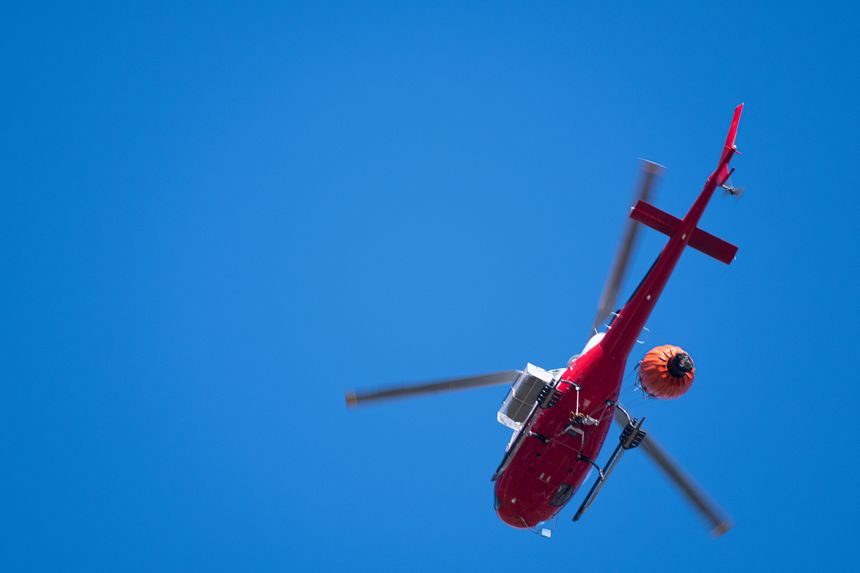 A red firefighting helicopter trailing a water bucket in a clear blue sky
