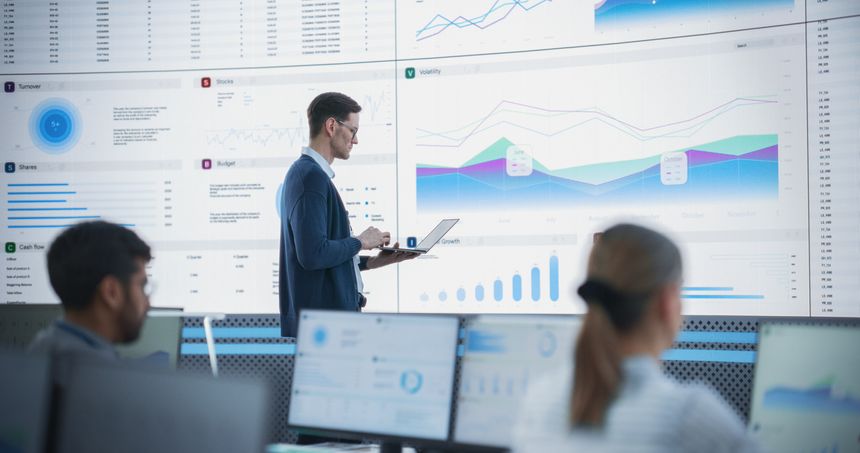 Male Senior Financial Analyst Reviewing Information And Reports on Big Digital Screen in Modern Monitoring Office. Diverse Employees Working Behind Desktop Computers In Investment Management Company.