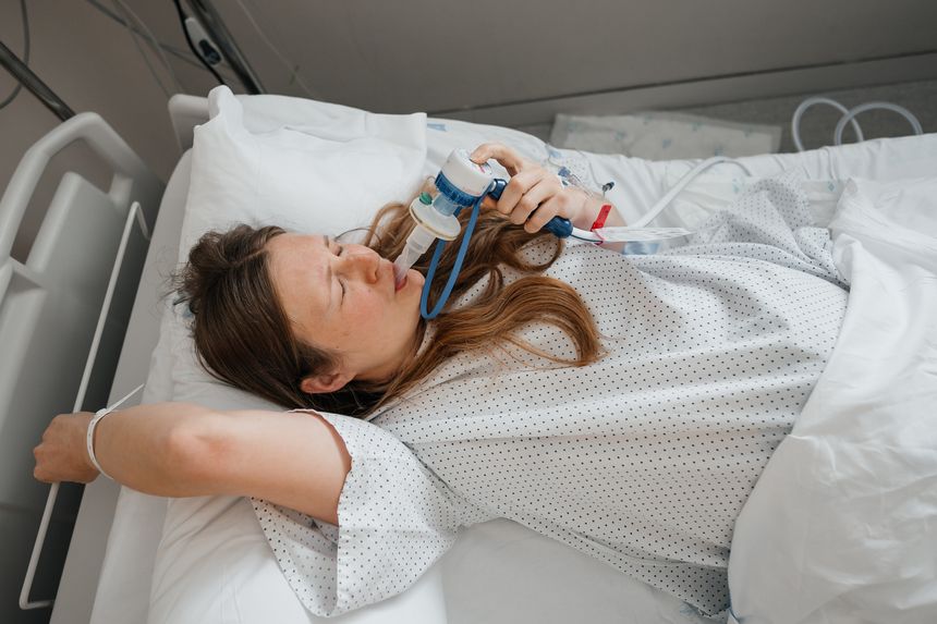 A pregnant woman lies in a hospital bed, gripping the bed rail and inhaling gas and air for pain relief while experiencing contractions during labor.