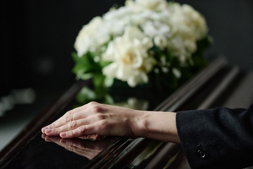 Caucasian middle aged woman resting hand on closed coffin during funeral ceremony, blurred bouquet of flowers in background, expressing grief and loss