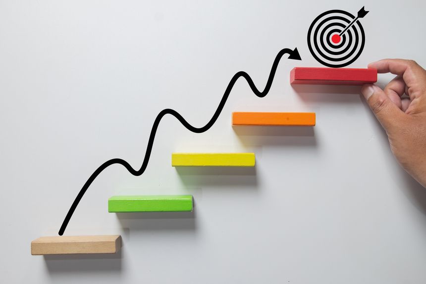 Business growth and success concept. Hand placing a wooden block on colorful stairs with an upward arrow leading to a target icon, symbolizing progress, achievement, and strategic planning.