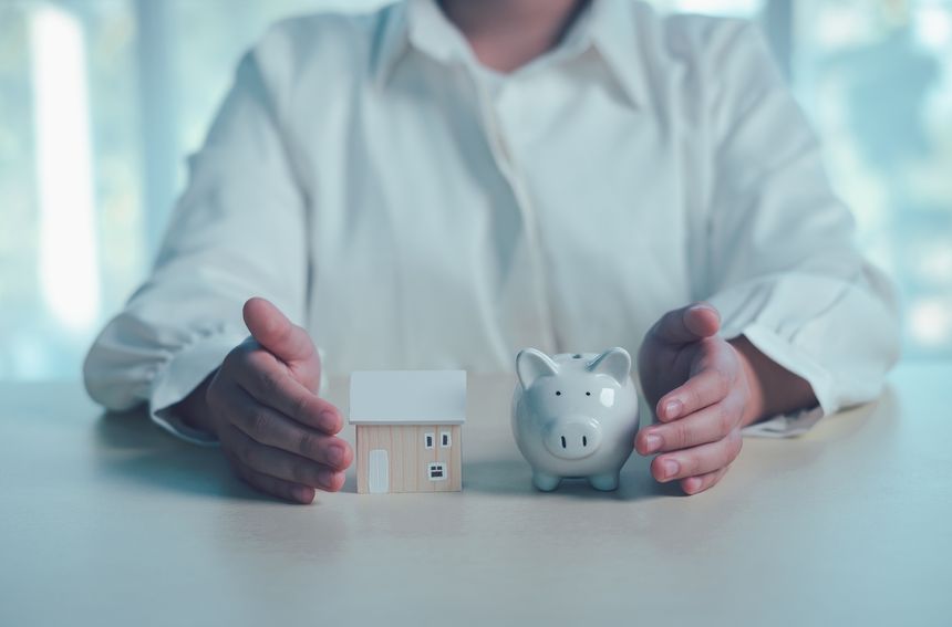 Home insurance and financial protection concept with hands protecting house model and piggy bank, representing property investment, savings planning, risk management and family financial security.