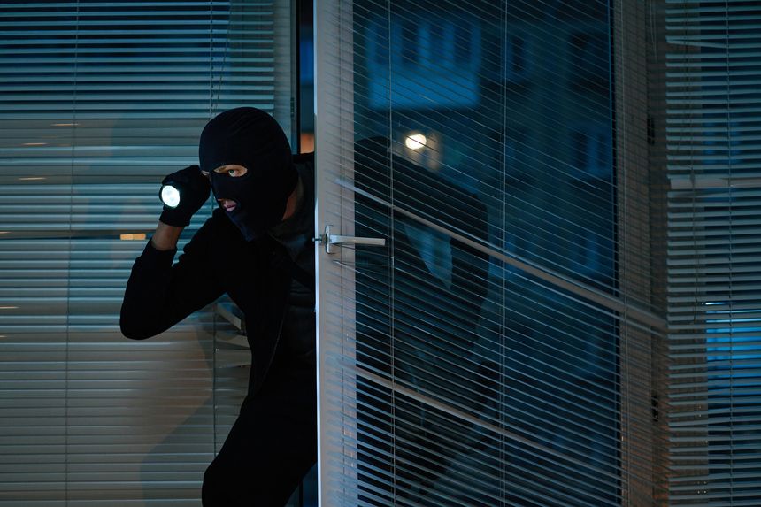 Man wearing black balaclava entering office at night holding flashlight, crouching near glass door, searching for valuables, engaging in theft activity