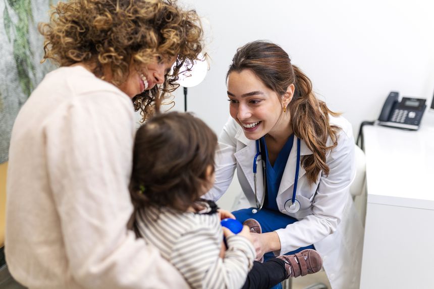 Pediatrician Examining Little Child. Friendly Female Doctor Smiling At Toddler During Medical Checkup. Healthcare And Medicine Concept. Pediatric Care For Baby With Mother In Hospital Office.