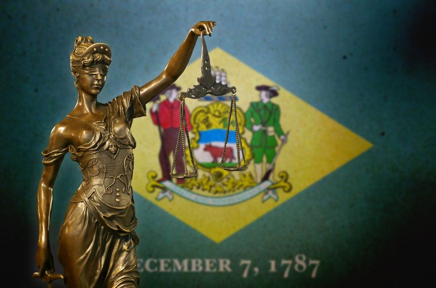 Close-up of a small bronze statuette of Lady Justice before a flag of Delaware.