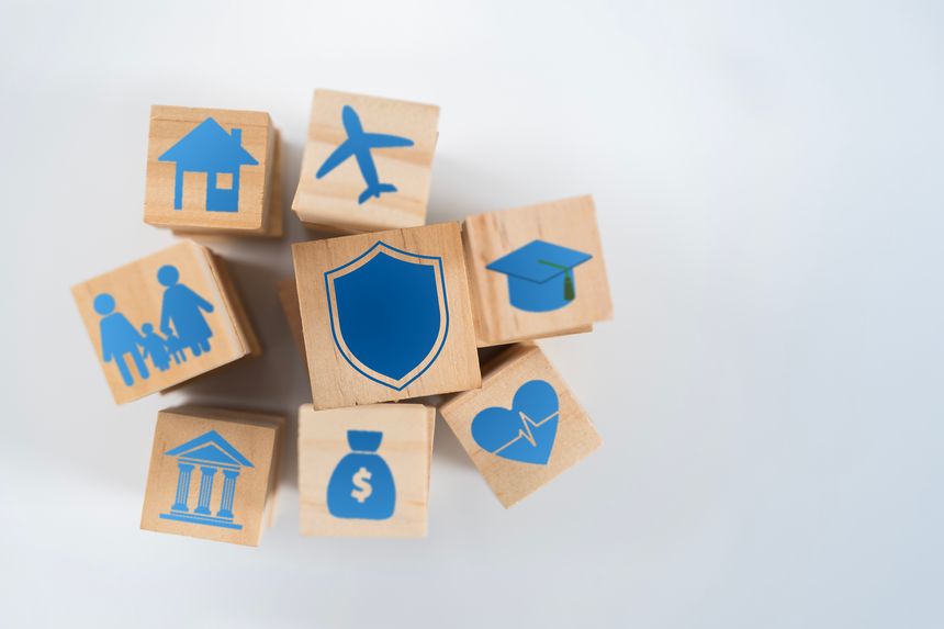 Wooden blocks with a central shield and lifestyle icons illustrate comprehensive insurance, financial protection, risk planning, family security, healthcare, education, and asset coverage.