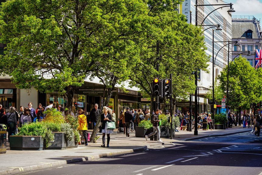 Oxford Street, London, United Kingdom – 17 April 2025: The John Lewis department store on Oxford Street displays its facade and shop windows on a sunny spring day. The scene captures everyday life and retail activity in central London during daytime. The shopfront is framed by green spring trees lining the pavement.