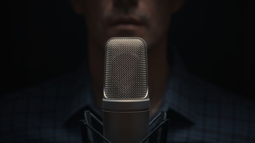 A professional studio microphone in dramatic low-key lighting with an out-of-focus man in the background. The image captures a clean, high-end audio production atmosphere, perfect for illustrating podcasts, voiceovers, narration, broadcasting, music recording, sound engineering, or professional content creation. Minimalistic composition draws attention to the mic's texture and premium build.