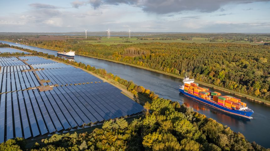 Container ship and car ferry navigate the Kiel Canal alongside vast solar panel fields and wind turbines, set against a backdrop of forest.
