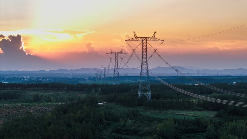 A beautiful sunset scene featuring high-voltage transmission towers standing in a vast field with greenery and distant mountains.