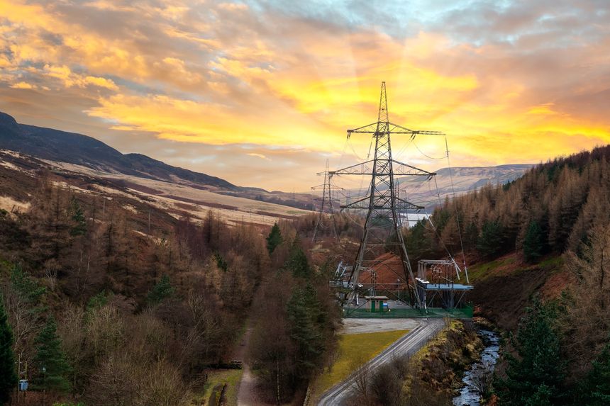 La escena captura una puesta de sol detrás de las montañas, con líneas eléctricas que se alzan imponentes. Los árboles bordean un sendero junto a un arroyo en el valle.