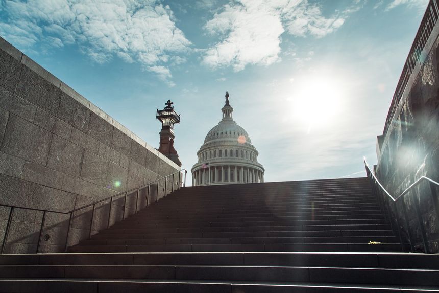 Visitors ascend the steps towards the iconic Capitol building in Washington DC, with sunlight illuminating the scene and clouds scattered above, capturing a moment of exploration and history.