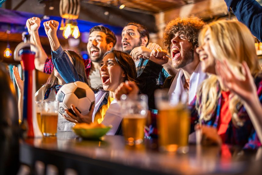 Fans celebrating victory with a soccer ball, beer and chips at bar counter. Young people shouting and cheering for their football team in a rustic pub interior.