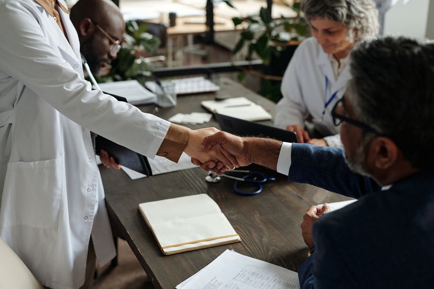 Caucasian woman in lab coat shaking hands with Black middle aged man in suit while two other professionals working at table with documents and laptop