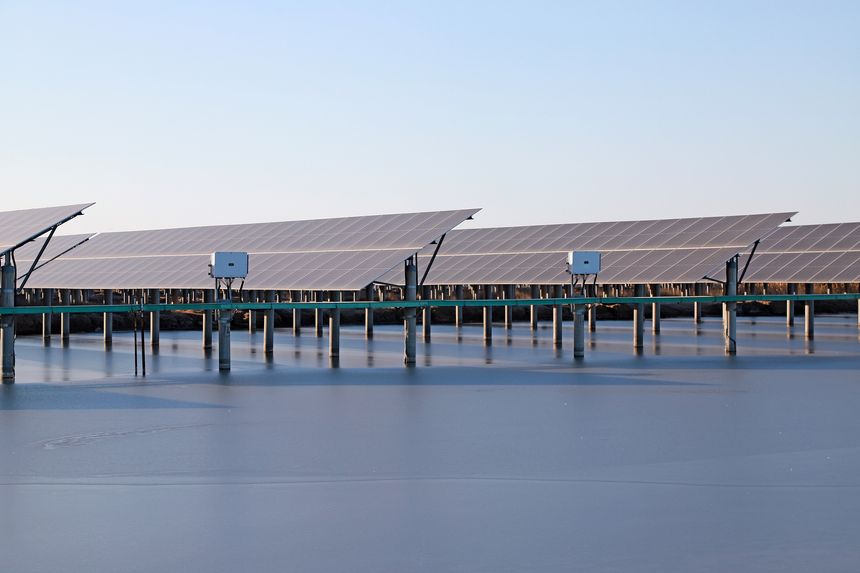 This photograph showcases an array of solar panels installed on a floating power station. Neatly arranged dark blue solar panels are mounted on concrete pillars and green supports above calm water, with white inverters suspended beneath. Against a light gray sky, it creates an industrial landscape that blends technology and ecological beauty, embodying the concept of integrating renewable energy development with the environment, making it ideal for visual content related to clean energy and sustainable development.