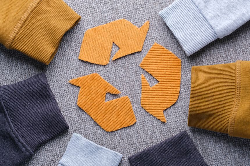 Sweater sleeves in shades of gray, mustard, and light blue surround a recycling symbol made from orange fabric. Concept of clothing recycling, sustainable fashion, and textile waste reduction. top view