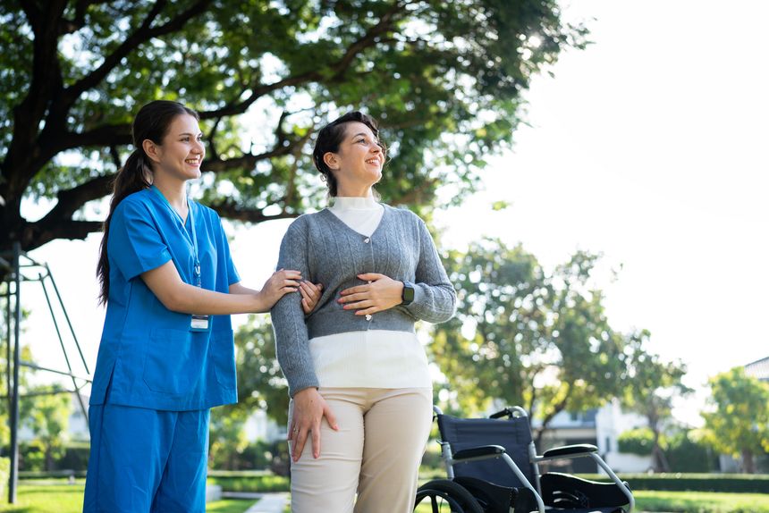 A nurse in blue scrubs helping a woman stand near a wheelchair in a sunny park, offering care and encouragement.