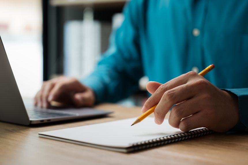 A man is writing on a notebook with a pencil. He is focused on his work and he is in a serious mood. The notebook is placed on a wooden desk, and there is a laptop nearby
