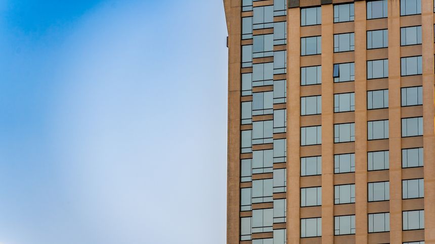 A partial view of a contemporary high-rise building featuring a grid of reflective windows and tan brick cladding. The architectural lines are sharp and geometric, set against a clear blue sky. The image captures modern urban design and vertical development.