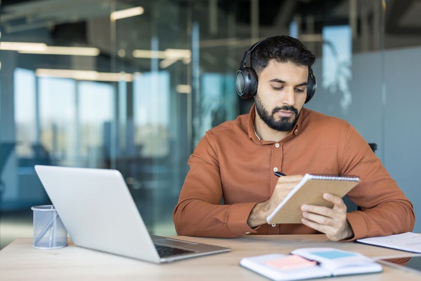 Young man with headphones works on a laptop in a modern glass-walled office, focused on taking notes in a notebook while studying or attending an online course for professional growth