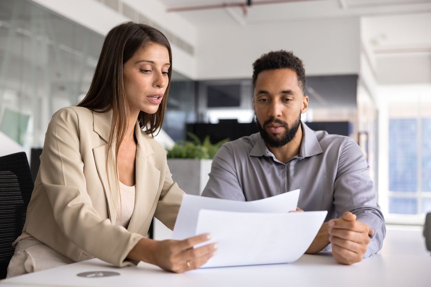 Serious young Latin business professional woman explaining paper to male African coworker, giving consultation about financial report, agreement text, legal document at work table