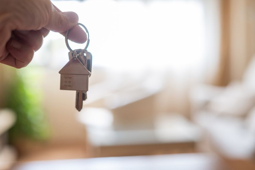 Hand holding house keys with house shaped keychain in front of blurred living room background, representing new home purchase, property investment, or rental agreement
