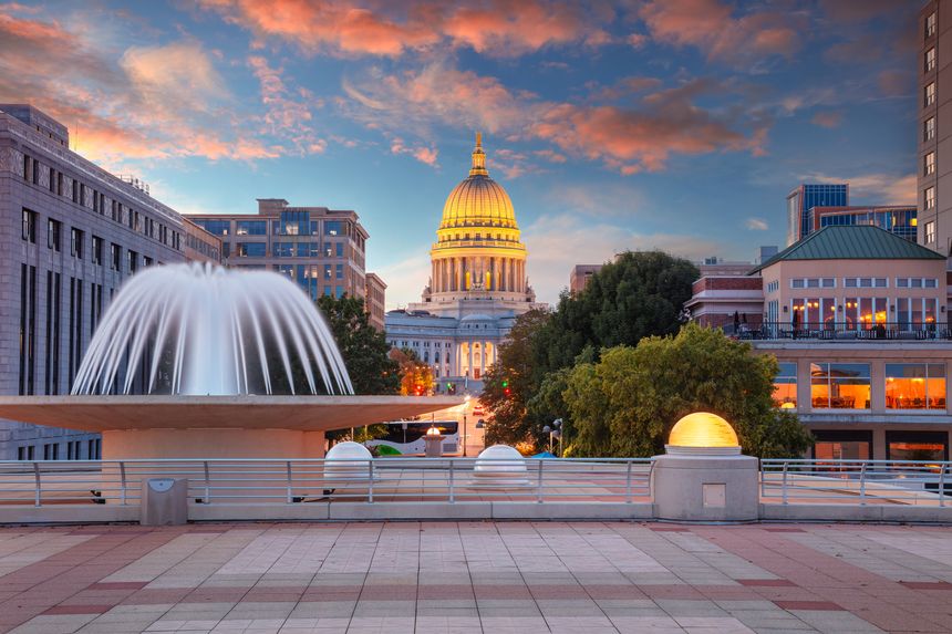 Madison, Wisconsin's Capitol building stands illuminated against a vivid autumn sunset. The scene features a prominent fountain in the foreground, with water cascading elegantly from a circular structure. Surrounding buildings display a mix of modern and classical architecture, with warm lights glowing from windows, adding to the serene ambiance. A wide, tiled plaza with railings and spherical lights enhances the composition. The sky is painted in hues of pink and orange, highlighting the stunning dome of the Capitol.