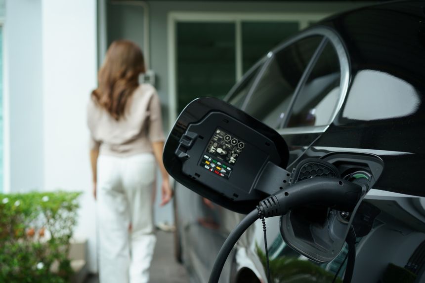 Close up of EV charging plug connected to black electric car at home, with Asian woman walking toward the house in blurred background. Renewable energy, sustainable lifestyle, and eco-friendly concept