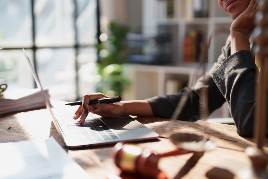 Legal professional at a wooden desk using a laptop and pen, reviewing documents with gavel and scales of justice nearby, focused on contracts and legal research