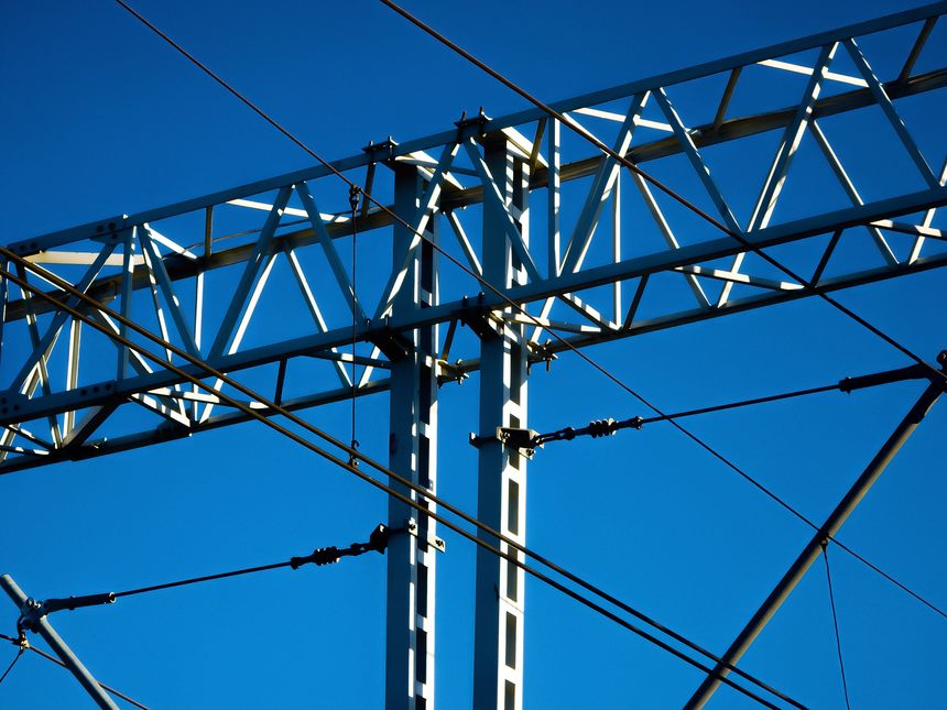 Close-up of metal overhead railway power gantry and wires against a clear deep blue sky, geometric steel framework and diagonal cables create strong lines