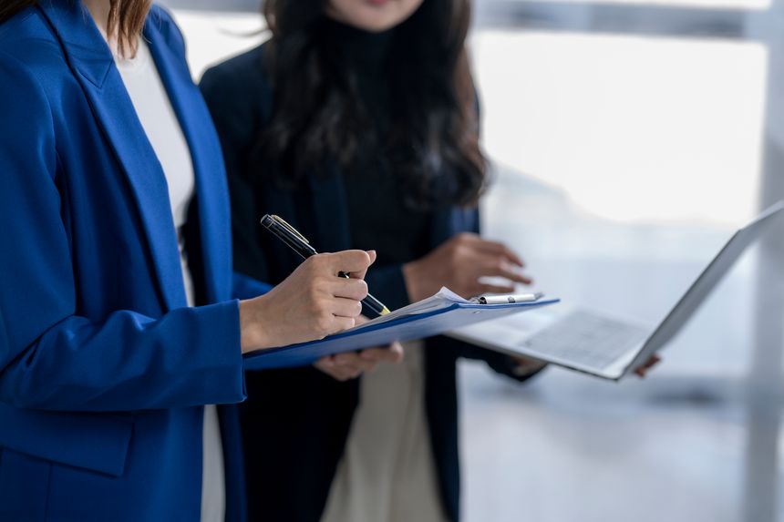 Businesswomen working together, reviewing a document on a clipboard and using a laptop