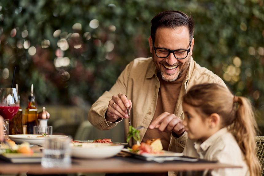 Father and young daughter share a joyful outdoor lunch at a restaurant patio, laughing and bonding over food; warm family moment ideal for lifestyle, parenting, hospitality, and marketing uses.