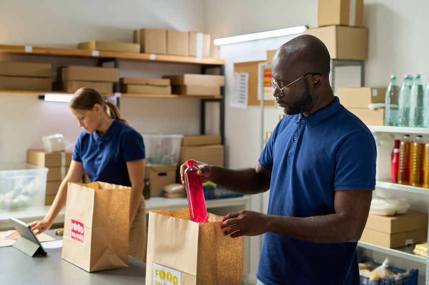 Black middle aged man packing food delivery bag with bottled drink while Caucasian young adult woman working on digital tablet in background, both preparing grocery orders in warehouse setting