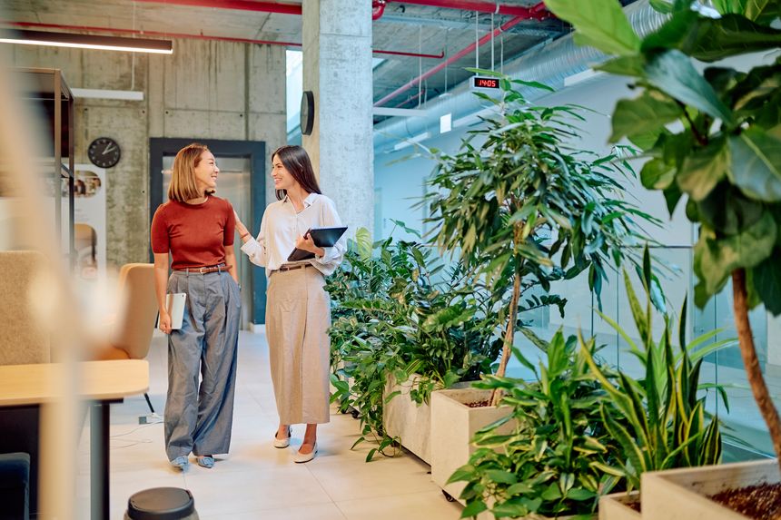 Two businesswomen walking through a vibrant green office, holding tablets and engaging in a lively discussion about their work projects