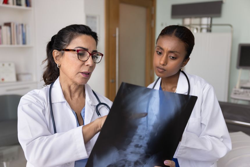 Two multiethnic female physicians, younger and older, study radiographic scans showing spinal condition, discuss diagnosis, evaluate treatment progress during medical consultation in examination room